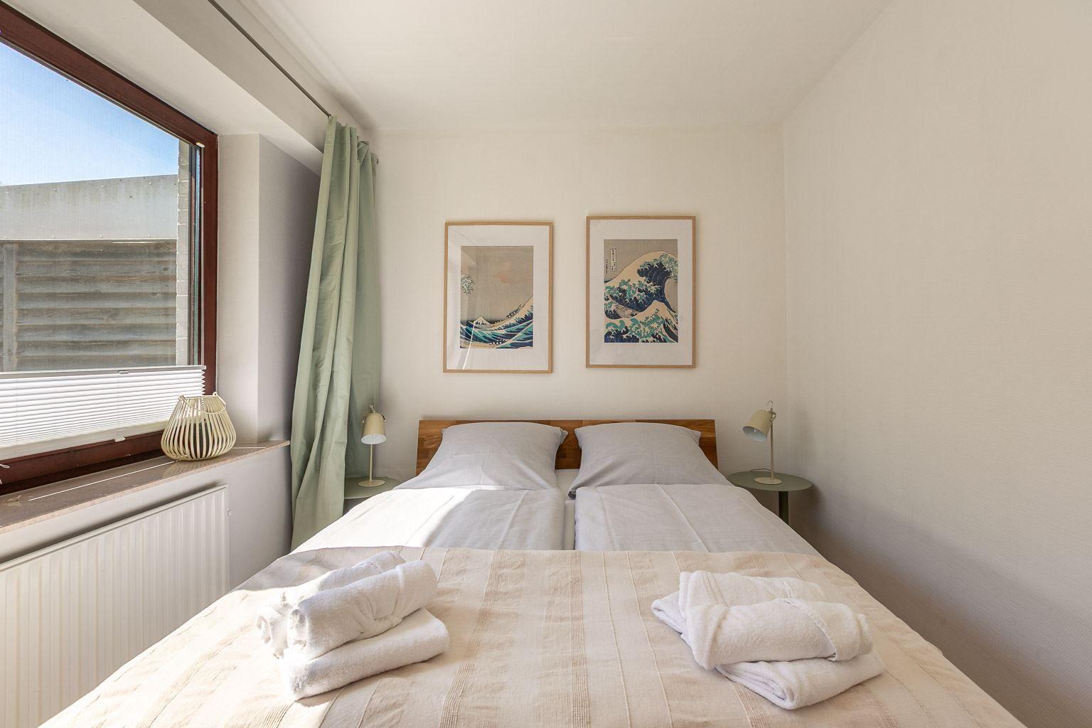 Schlafzimmer mit Doppelbett, zwei Nachttischen und Wandbildern. Fenster mit Jalousie und Vorhang.