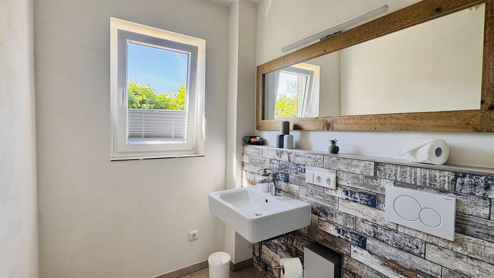 Badezimmer mit Waschbecken, Spiegel und Fenster mit Blick auf Grün.