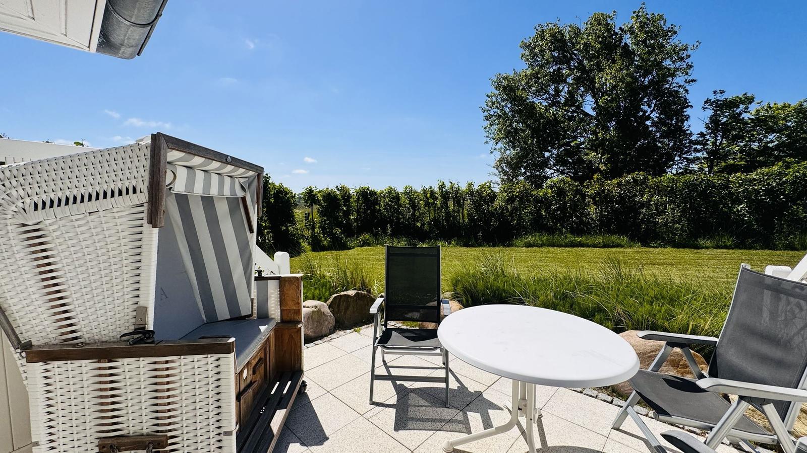 Terrasse mit Liegestuhl, Tisch und Strandkorb. Grünfläche und Bäume im Hintergrund.