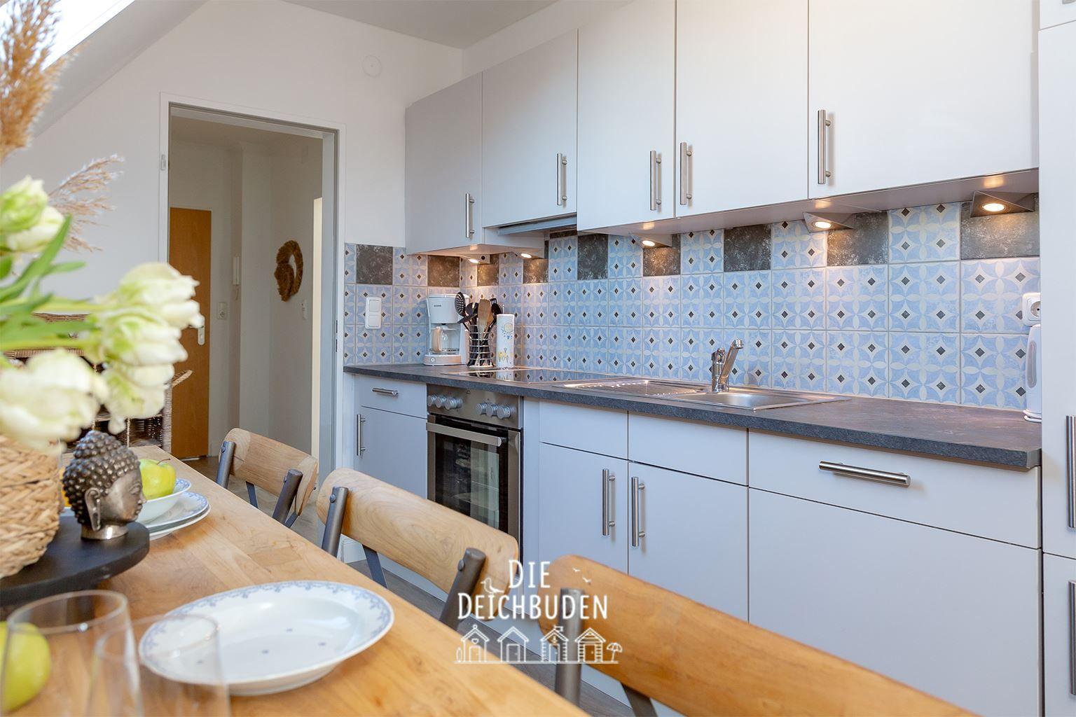 Küche mit weißen Schränken, blau gemusterter Wandfliese und Holztisch mit Stühlen.
