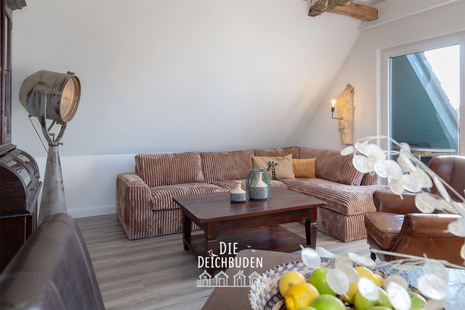 Wohnzimmer mit Ecksofa, Holztisch und Fenster. Deckenbalken und Stehlampe sichtbar.