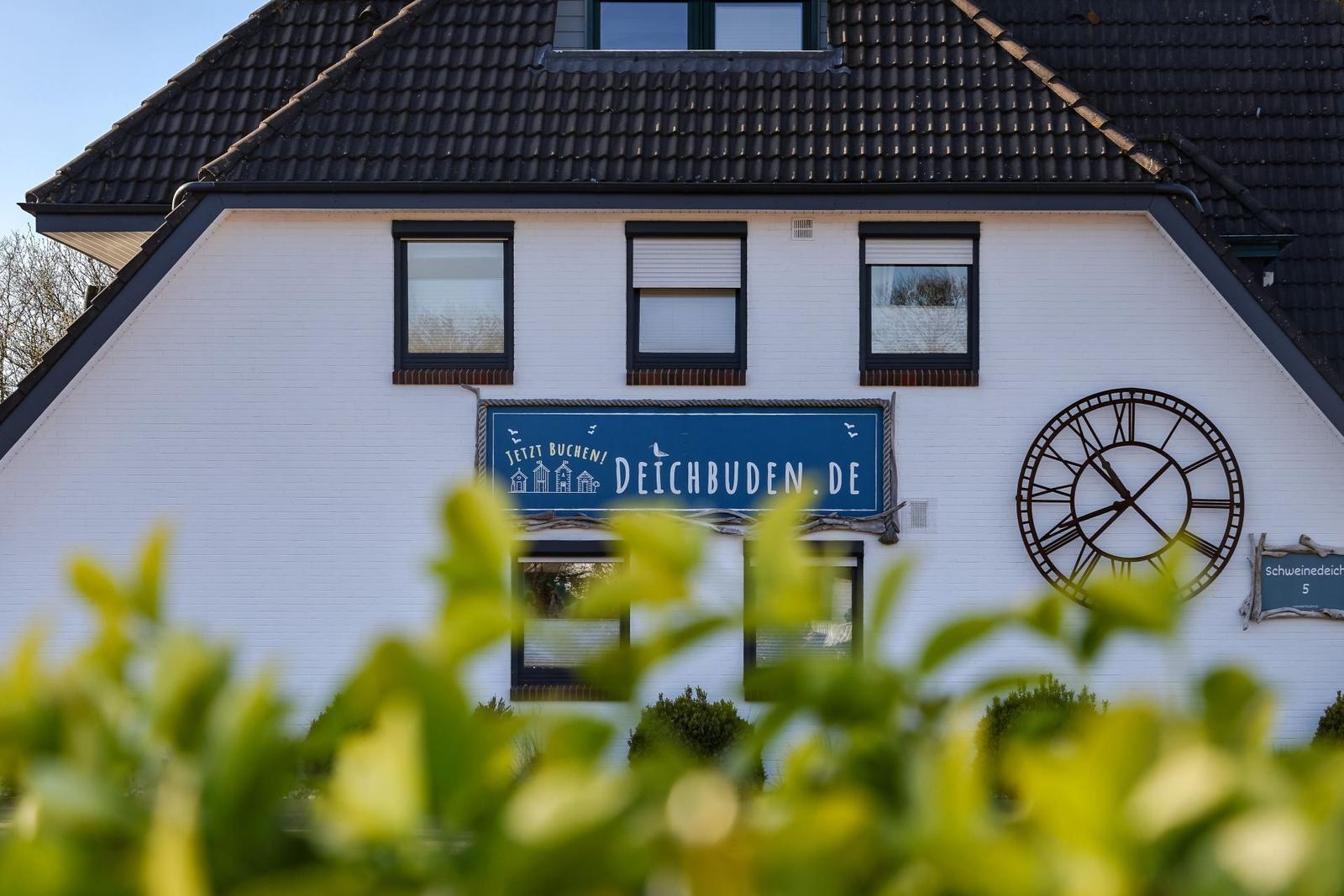 Weißes Haus mit dunklem Dach, Schild und großem Uhrwerk an der Fassade.