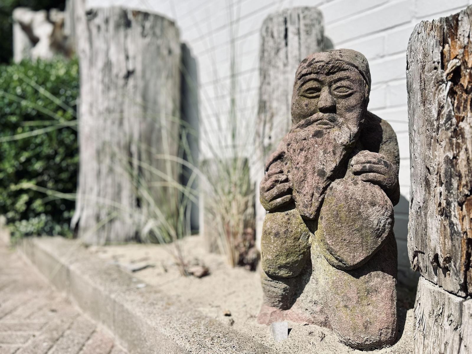 Steinskulptur eines Mannes mit Bart, umgeben von Holzstümpfen und Gras.