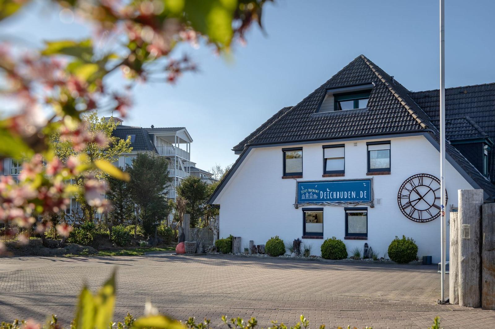 Weißes Haus mit dunklem Dach und großem Wanduhr. Vorhof mit gepflastertem Platz und Blumen.