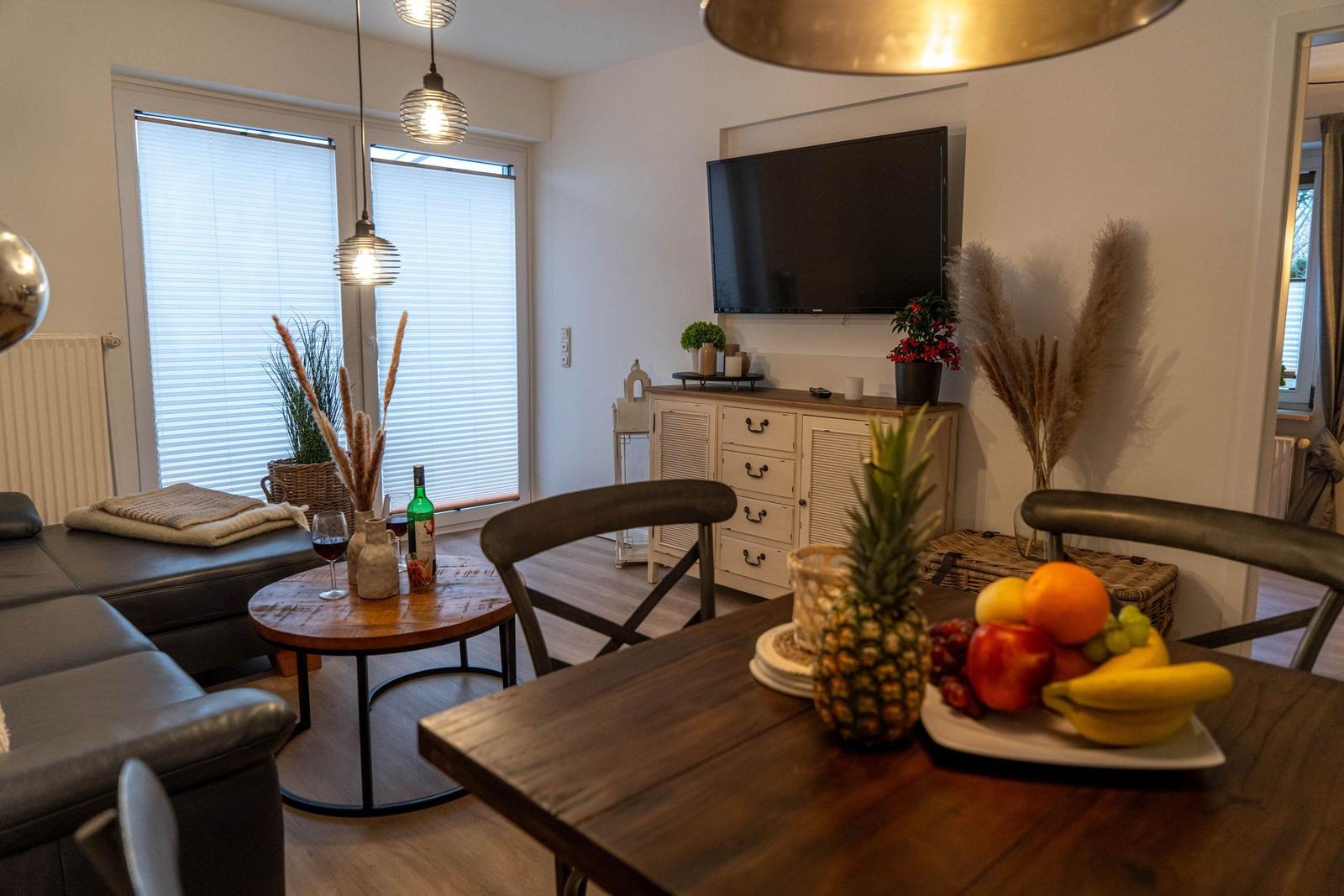 Essbereich mit Früchten und Wohnzimmer mit TV und Fenstern.