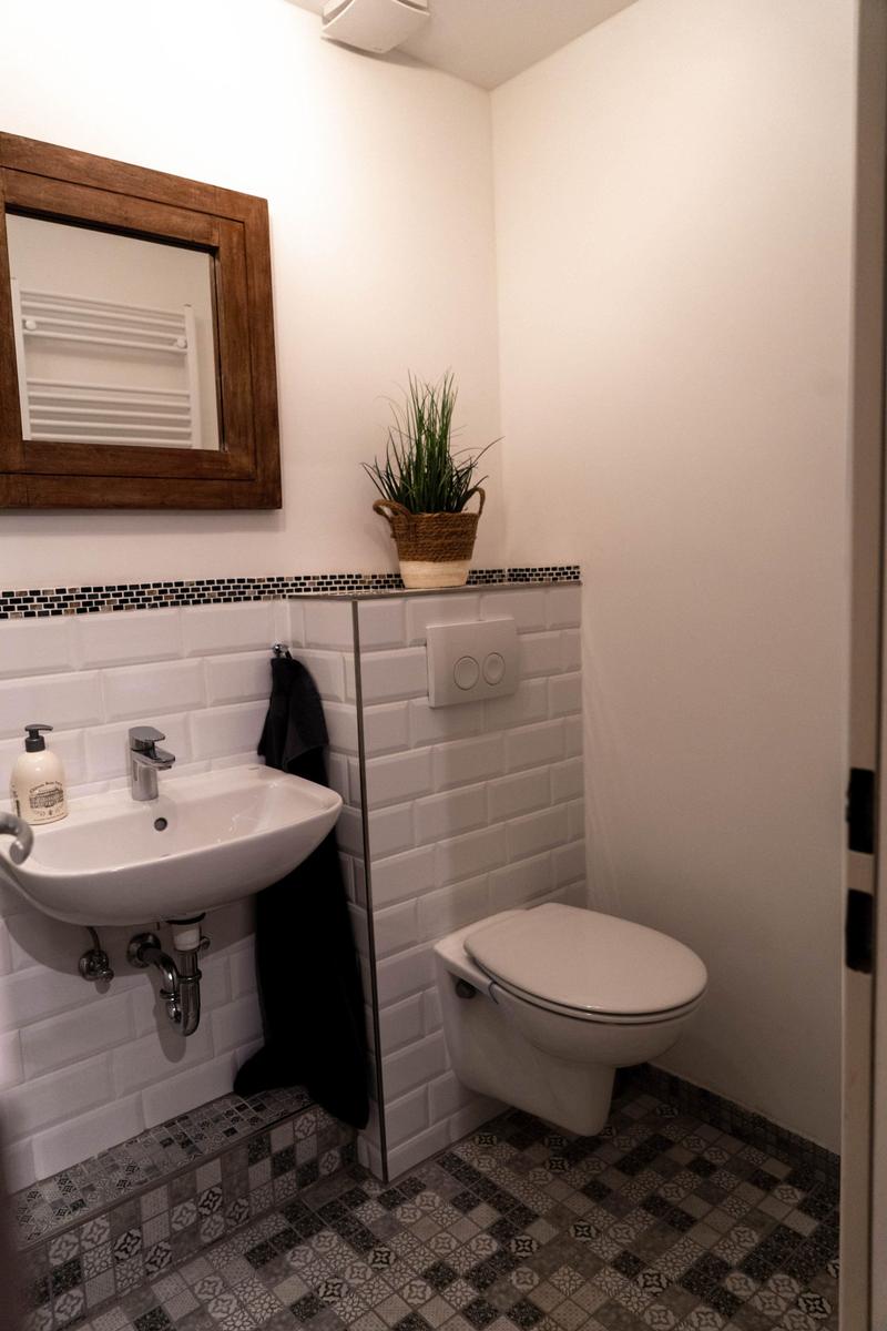 Badezimmer mit Waschbecken, Toilette und Pflanze. Fliesenboden und Spiegel mit Holzrahmen.