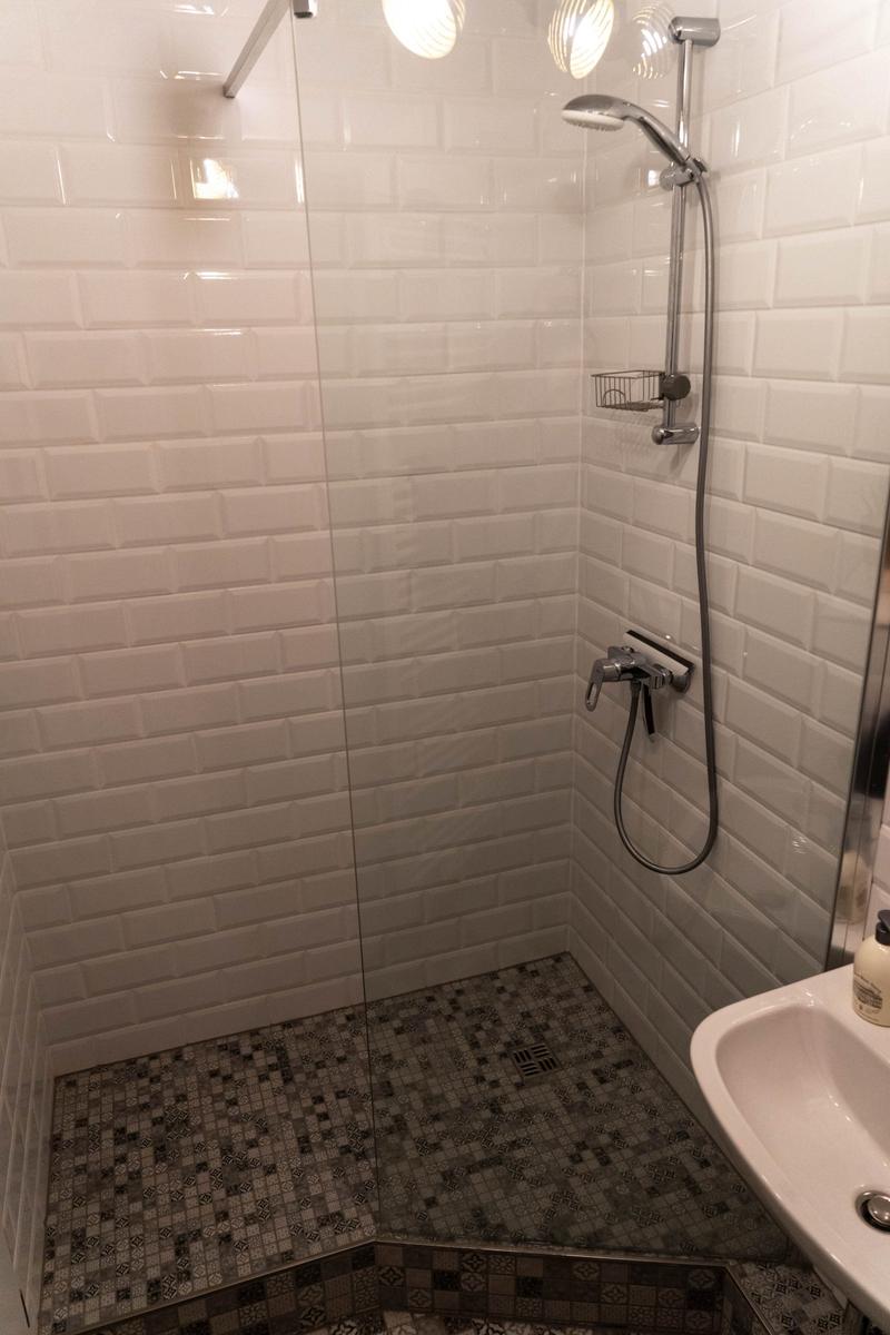 Dusche mit Glaswand, weißen Fliesen und Mosaikboden. Handbrause und Duschstange sichtbar.