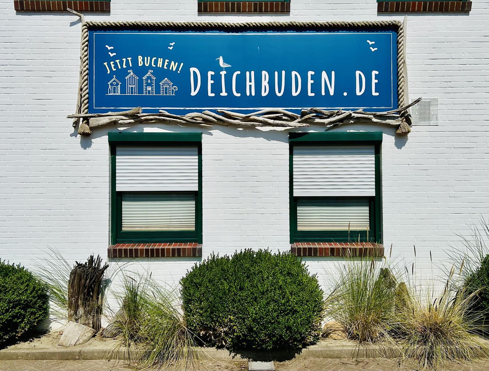 Weiß gestrichene Wand mit blauem Schild und zwei Fenstern mit Rollläden.