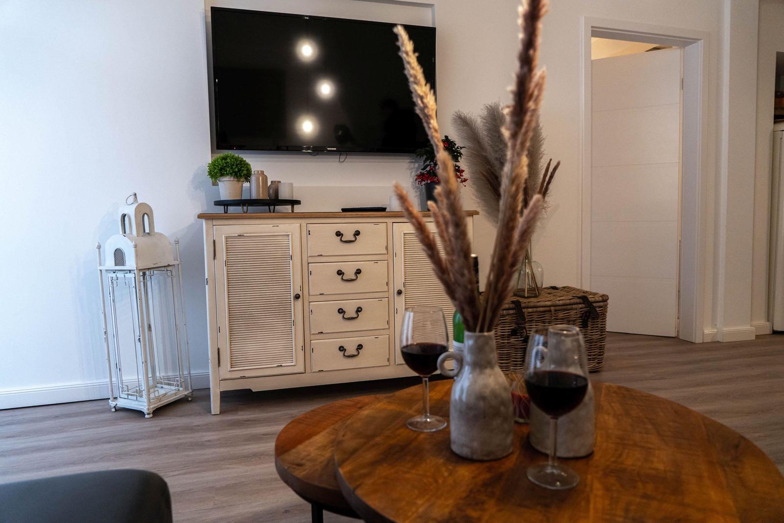 Rundes Holztisch mit zwei Weingläsern und Vasen mit Pampasgras. Hintergrund: Schrank und Wandfernseher.