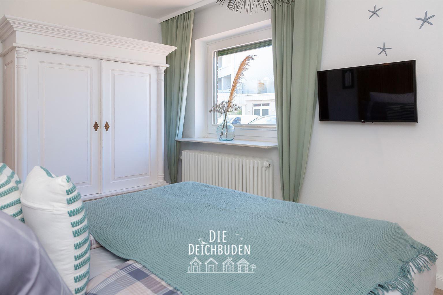 Schlafzimmer mit Bett, weiße Kommode, Fenster mit Vorhängen und Wand-TV.