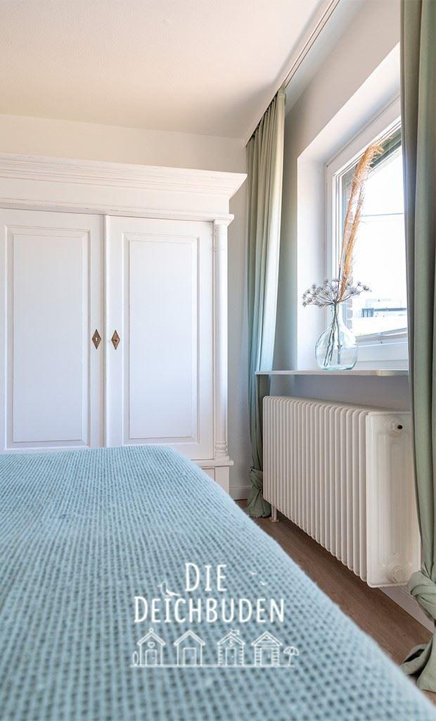 Schlafzimmer mit weißem Schrank, Fenster mit Vorhängen und Heizkörper.
