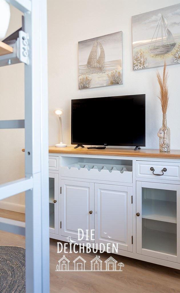 TV auf Holzboard über weißem Schrank mit Weinregal und Wandbildern von Segelbooten.