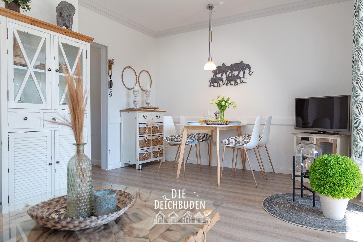 Ein Essbereich mit Tisch, Stühlen und Wanddekoration. Fernseher und Pflanzen sind ebenfalls sichtbar.