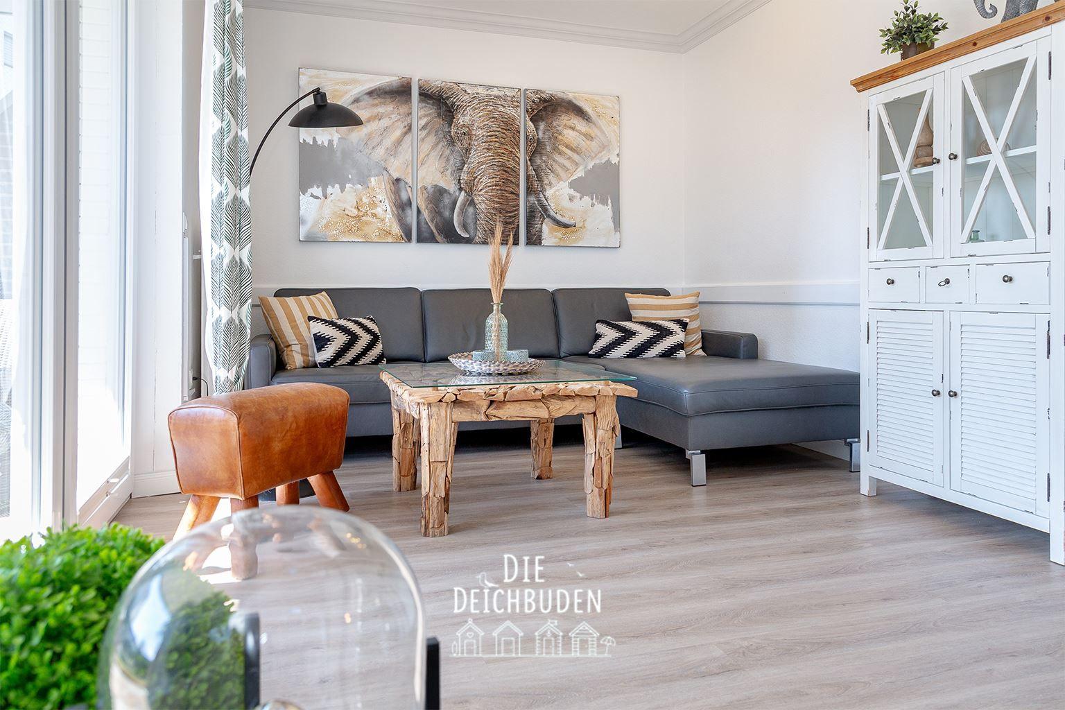 Ein Wohnzimmer mit grauem Sofa, Holztisch und weißem Schrank. An der Wand hängt ein Elefantenbild.