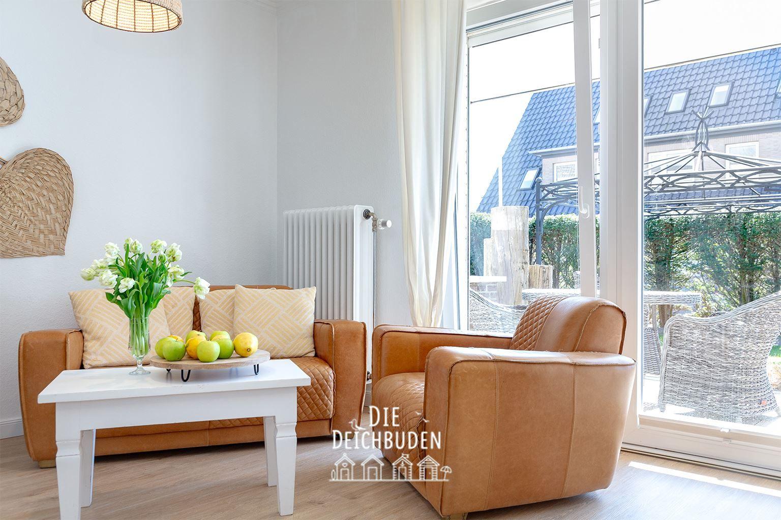 Lichter Raum mit braunen Ledersofa und Sessel. Weiße Tischplatte mit Blumen und Obst. Große Fenster zur Terrasse.