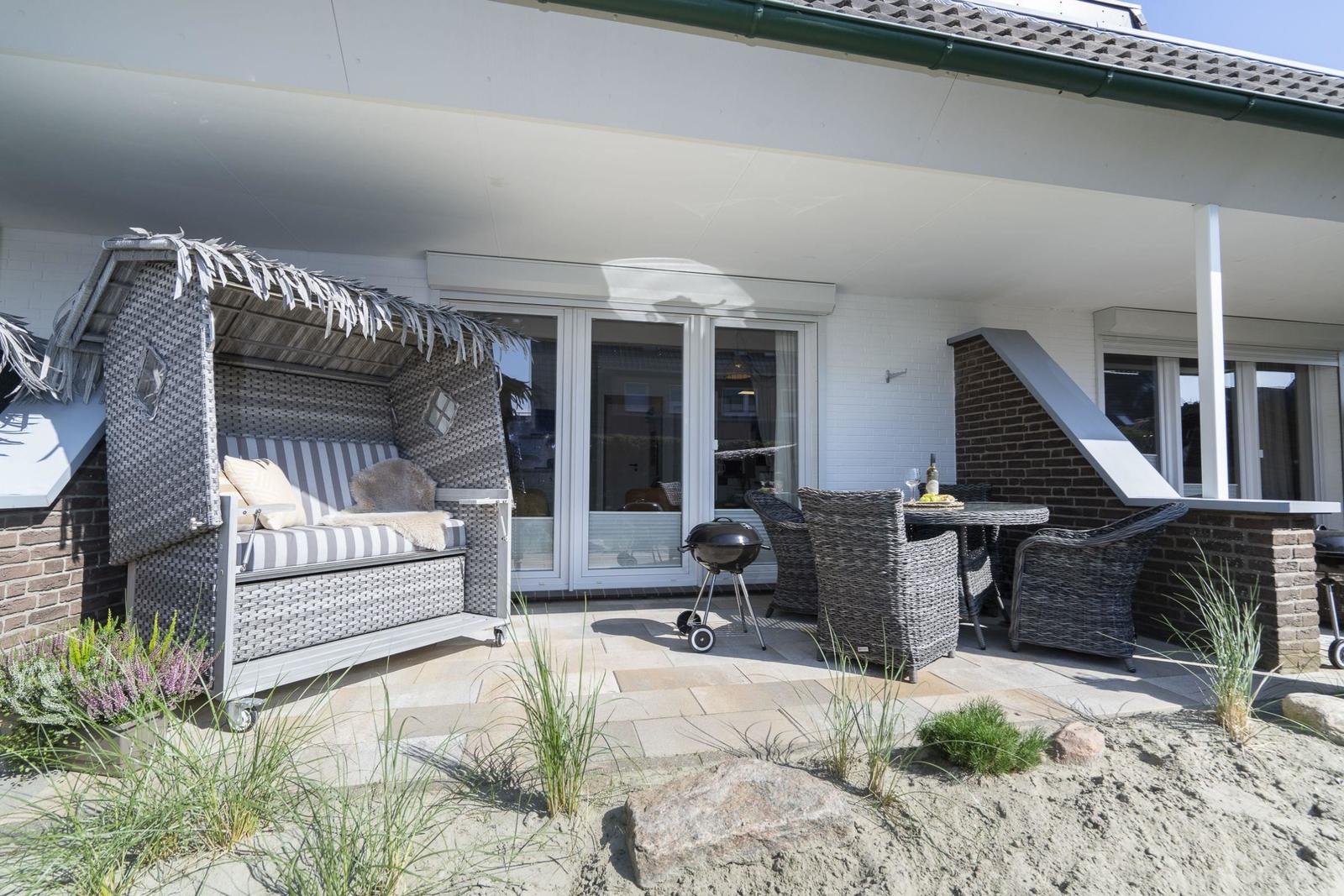 Terrasse mit Strandkorb, Grill und Sitzgruppe. Sand und Gras im Vordergrund.