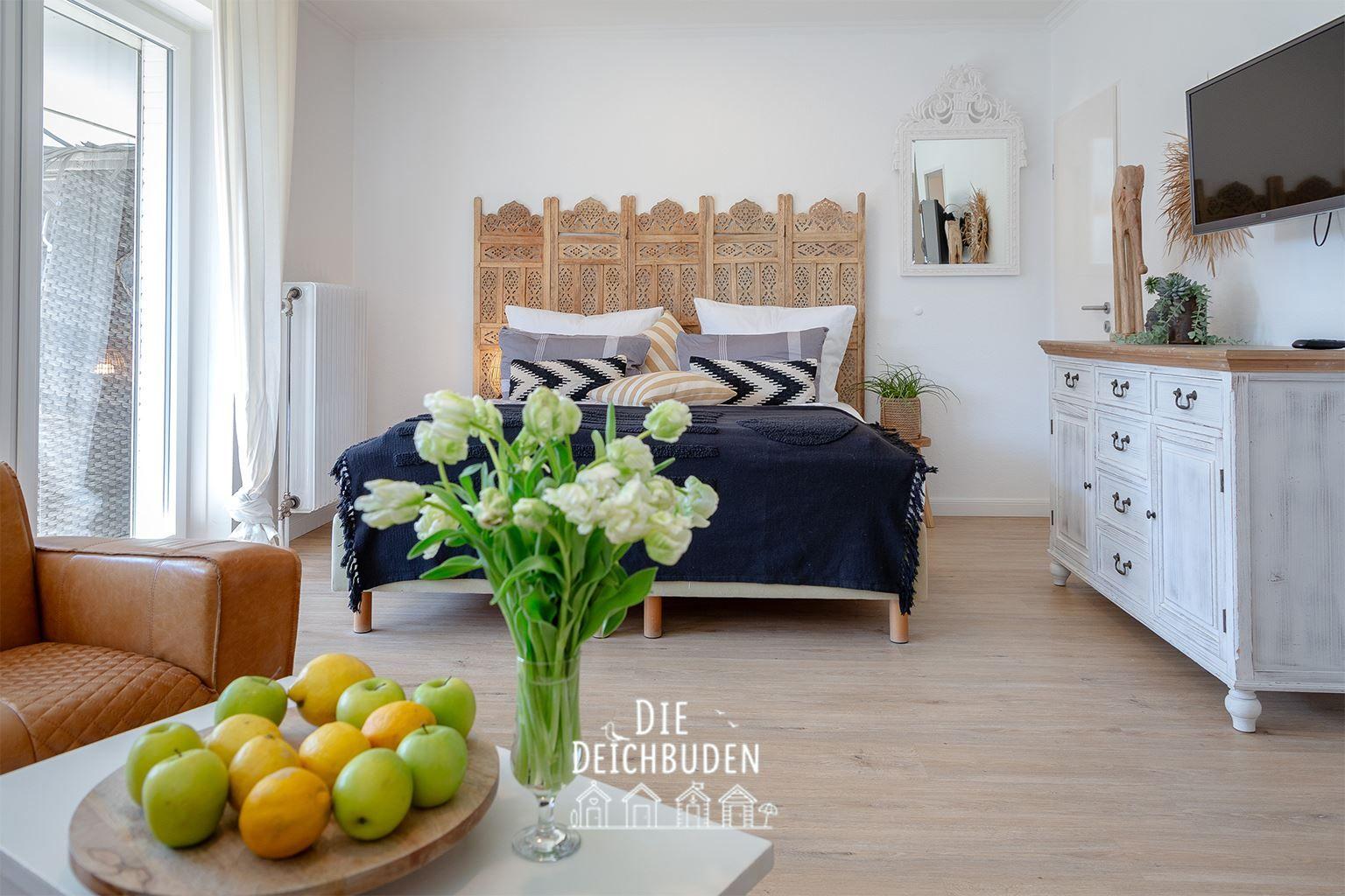 Schlafzimmer mit Bett, Schrank und Fenster. Auf Tisch Obst und Blumen.