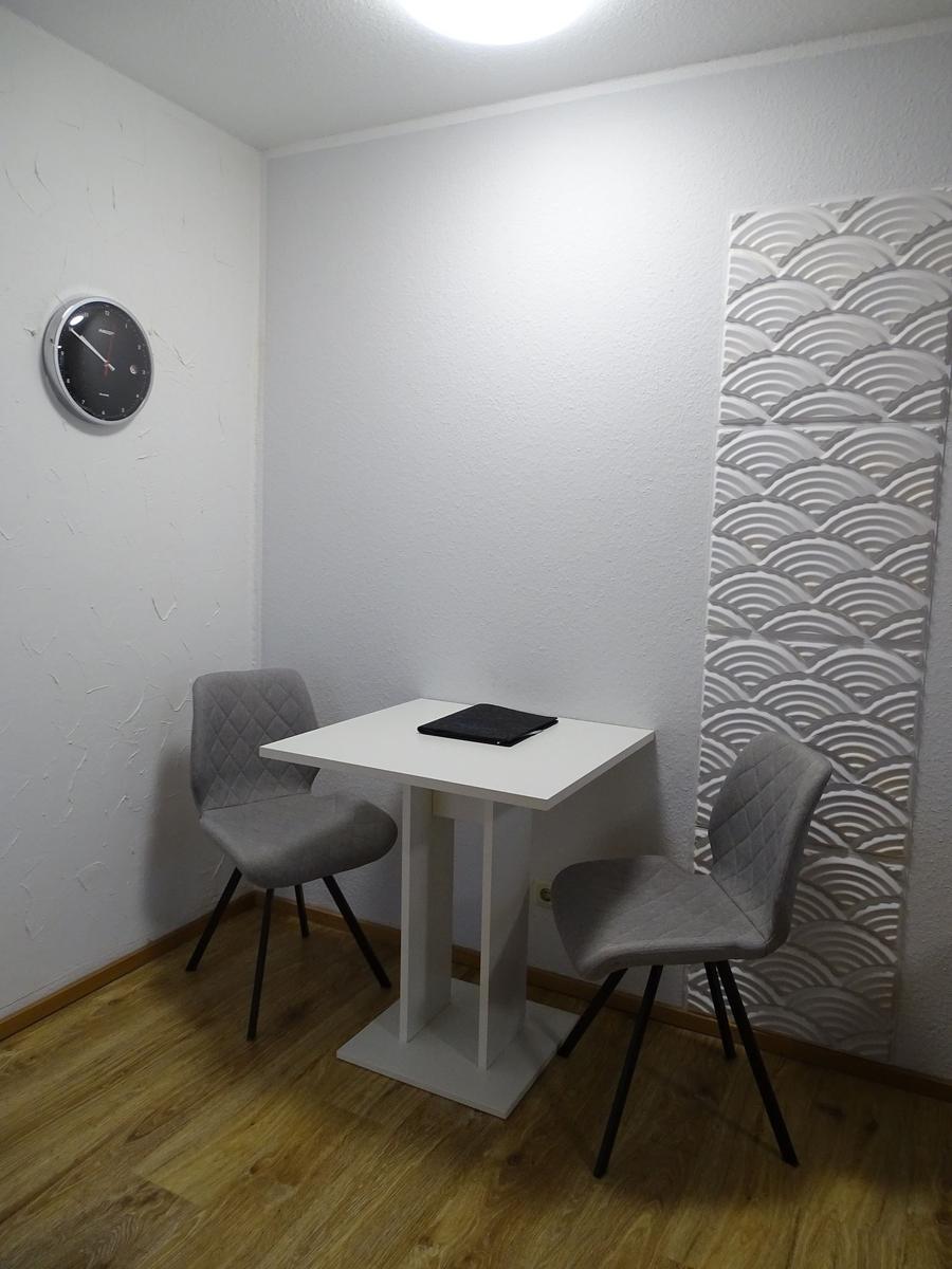 Ein Raum mit weißem Tisch, zwei grauen Stühlen und einer Wanduhr an der Wand.