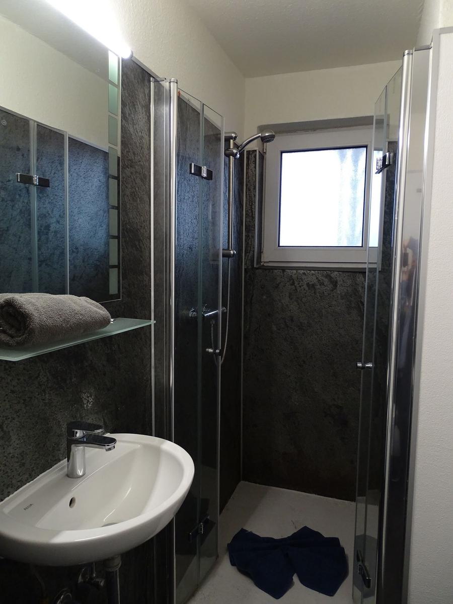 Badezimmer mit Dusche, Waschbecken und Spiegel. Dunkle Wandfliesen und Fenster.