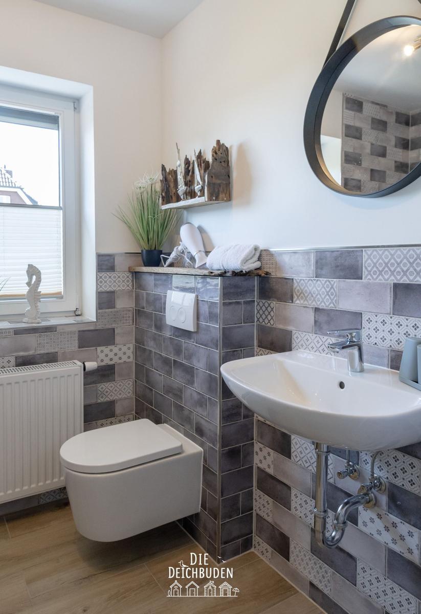 Badezimmer mit Toilette, Waschbecken und Fenster. Wandverkleidung aus grauen Fliesen.