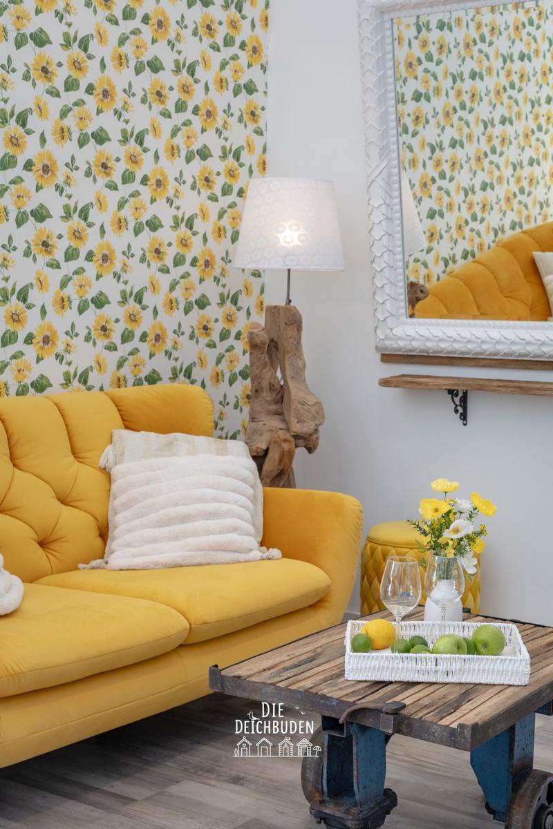 Gelber Sofa mit Kissen, Sonnenblumenwand, Holztisch und Spiegel.