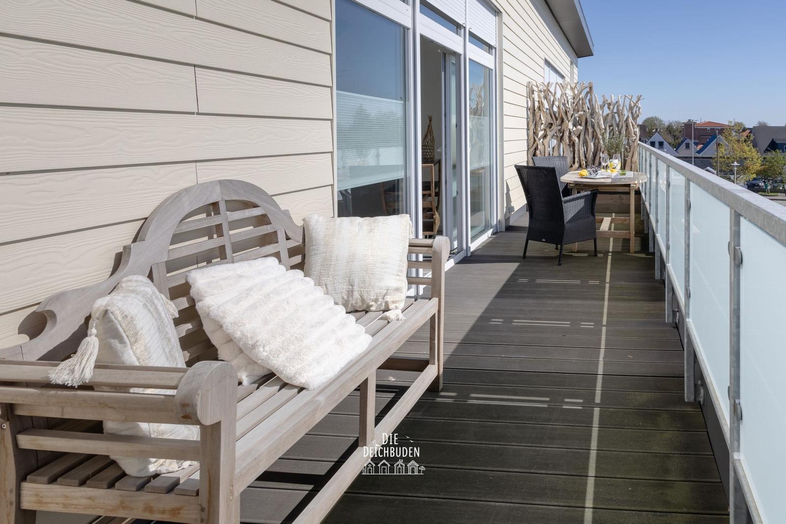 Terrasse mit Holzbank, Kissen und Tisch mit Stühlen. Glasgeländer und Hauswand im Hintergrund.