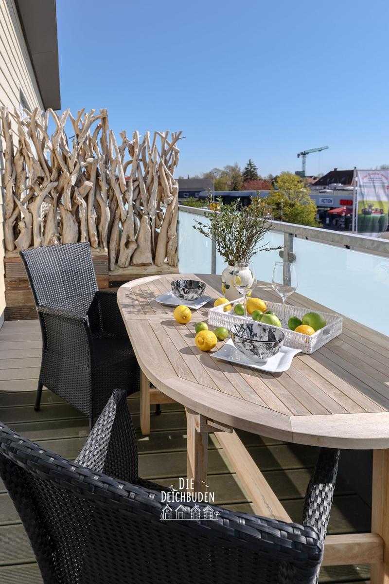 Terrasse mit Tisch, Stühlen und Obst. Hintergrund: Holzgeländer und Stadtansicht.