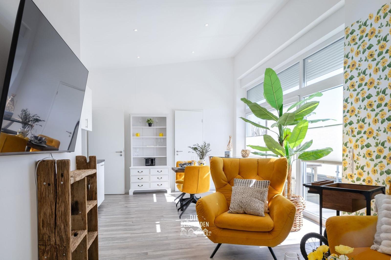 Wohnzimmer mit gelbem Sessel, Sonnenblumenwand und großen Fenstern.