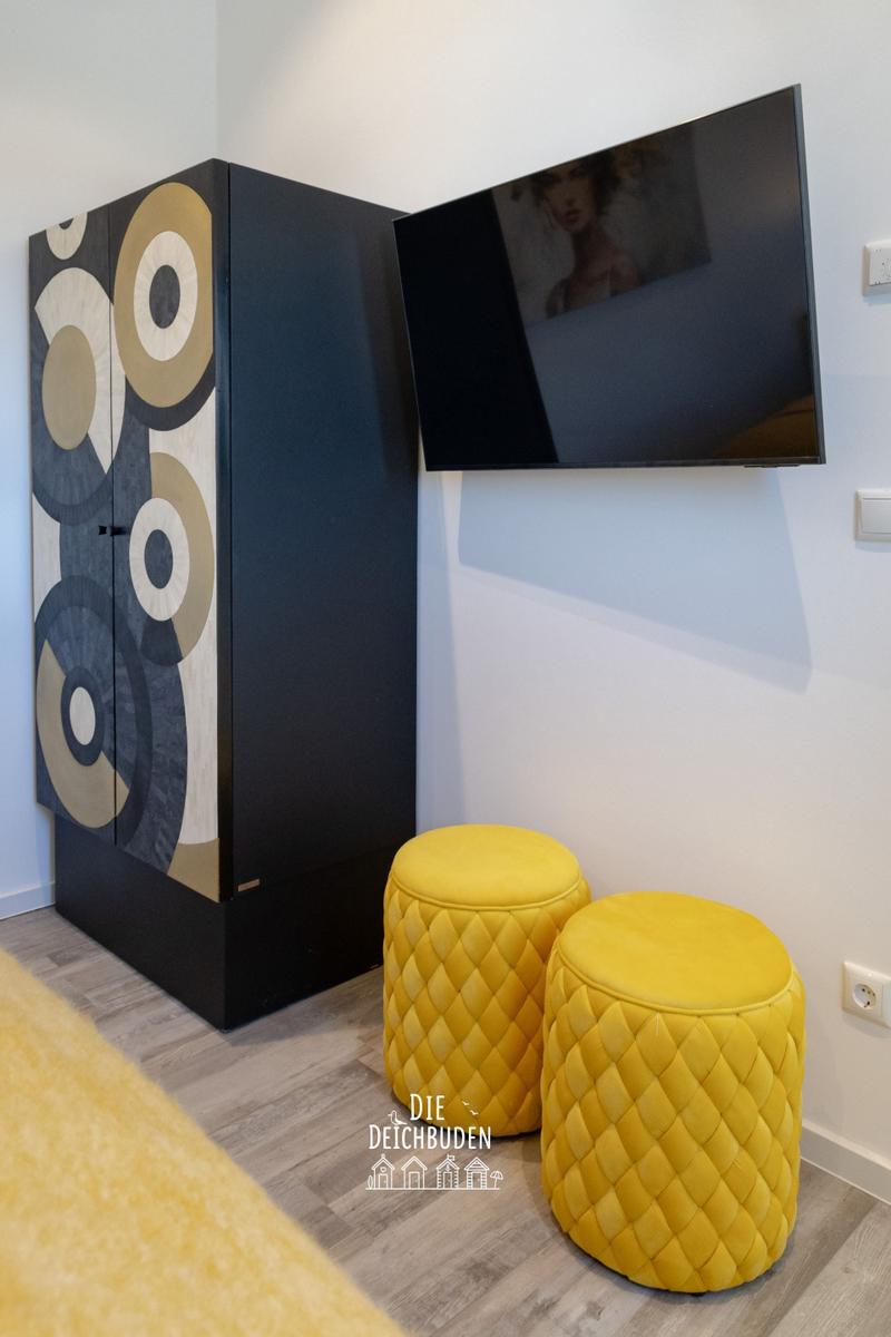 Schwarzer Schrank mit geometrischem Muster, zwei gelbe Hocker und Wand-TV.