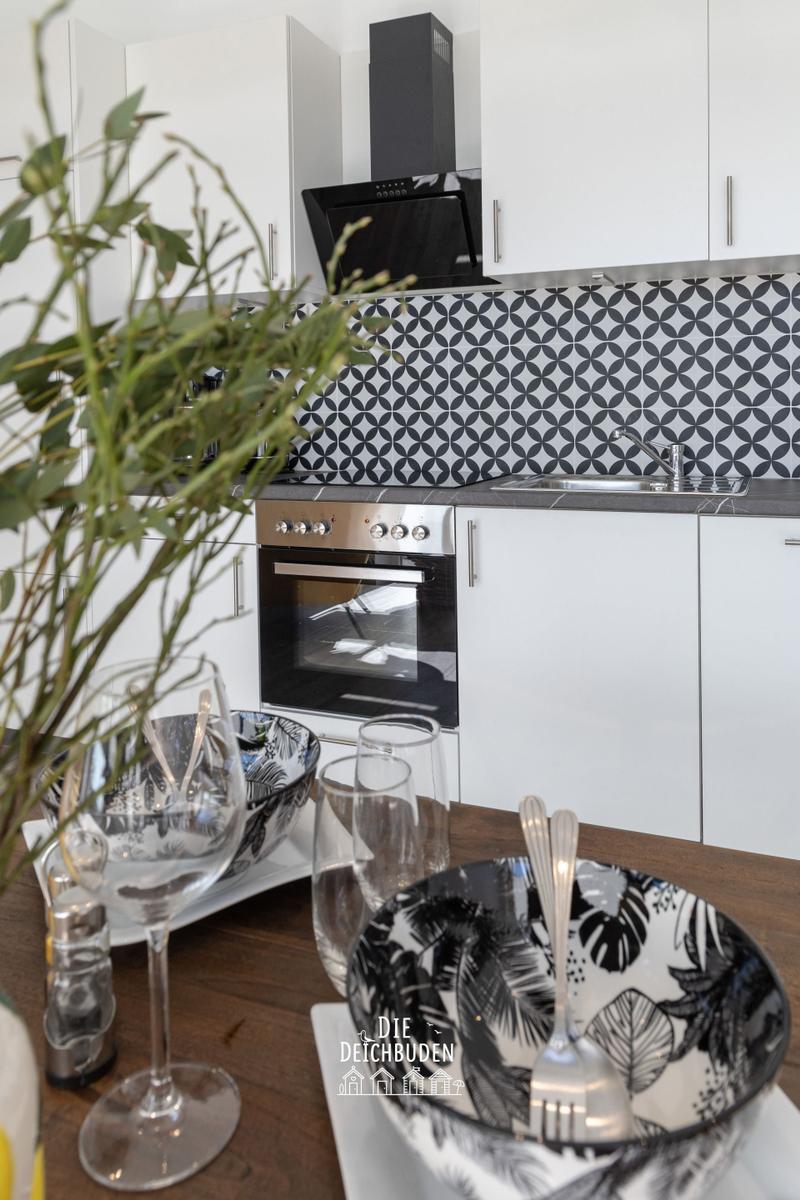 Küche mit weißen Schränken, Mosaikwand und Edelstahlgeräten. Tisch mit Geschirr und Pflanze im Vordergrund.