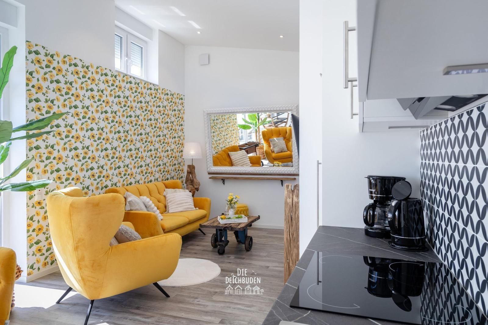 Wohnbereich mit gelben Sitzmöbeln und Sonnenblumenwand. Küche mit schwarzer Arbeitsplatte.