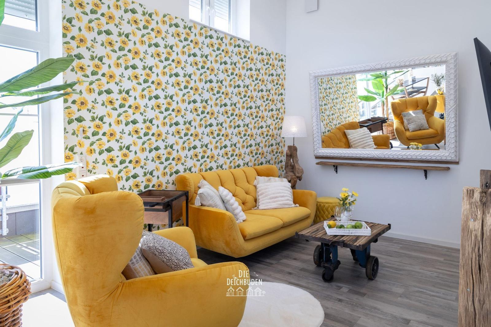 Wohnzimmer mit gelben Sitzmöbeln und Sonnenblumenwand. Spiegel, Holztisch und Pflanzen.