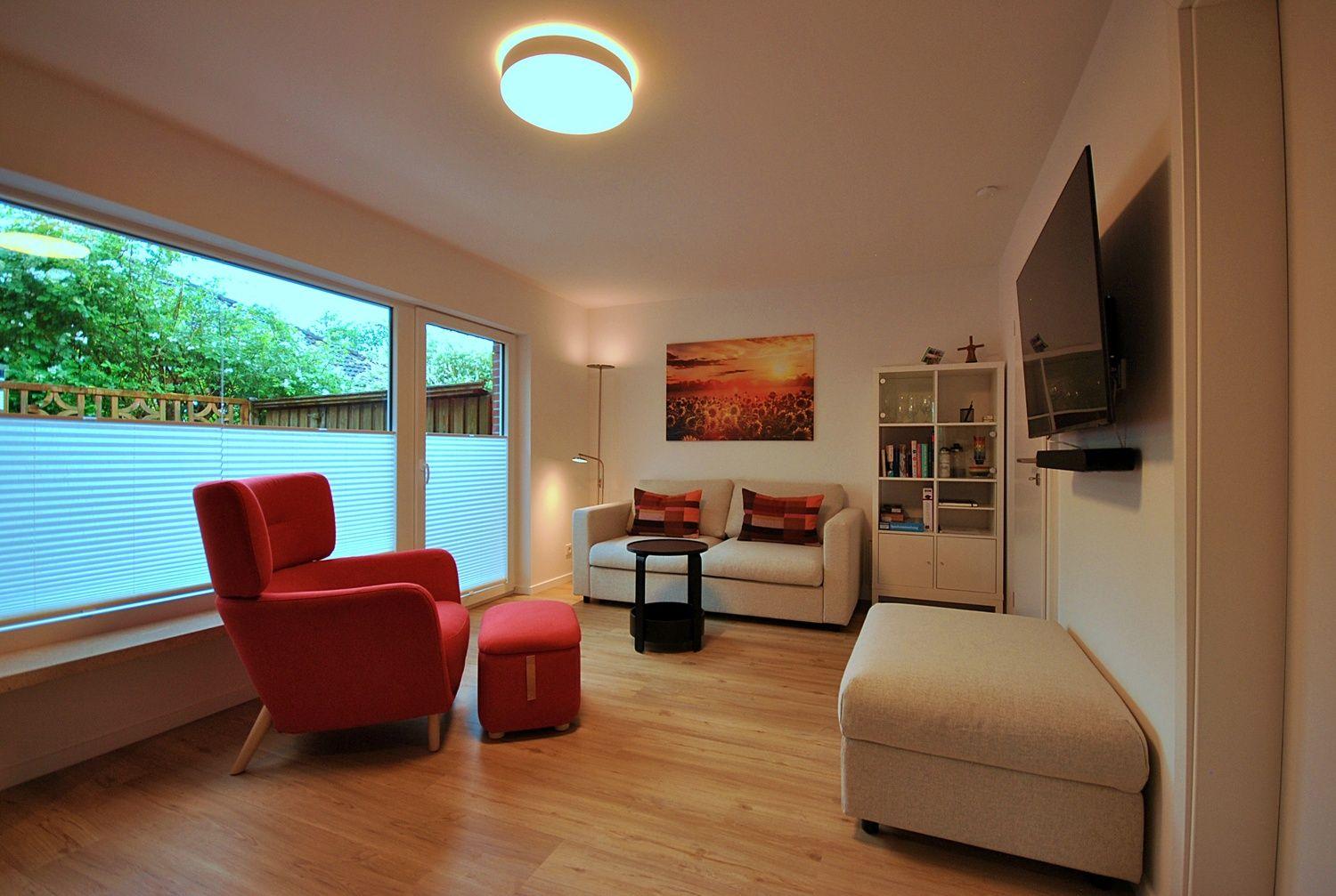 Wohnzimmer mit rotem Sessel, Sofa, Fernseher und großen Fenstern.