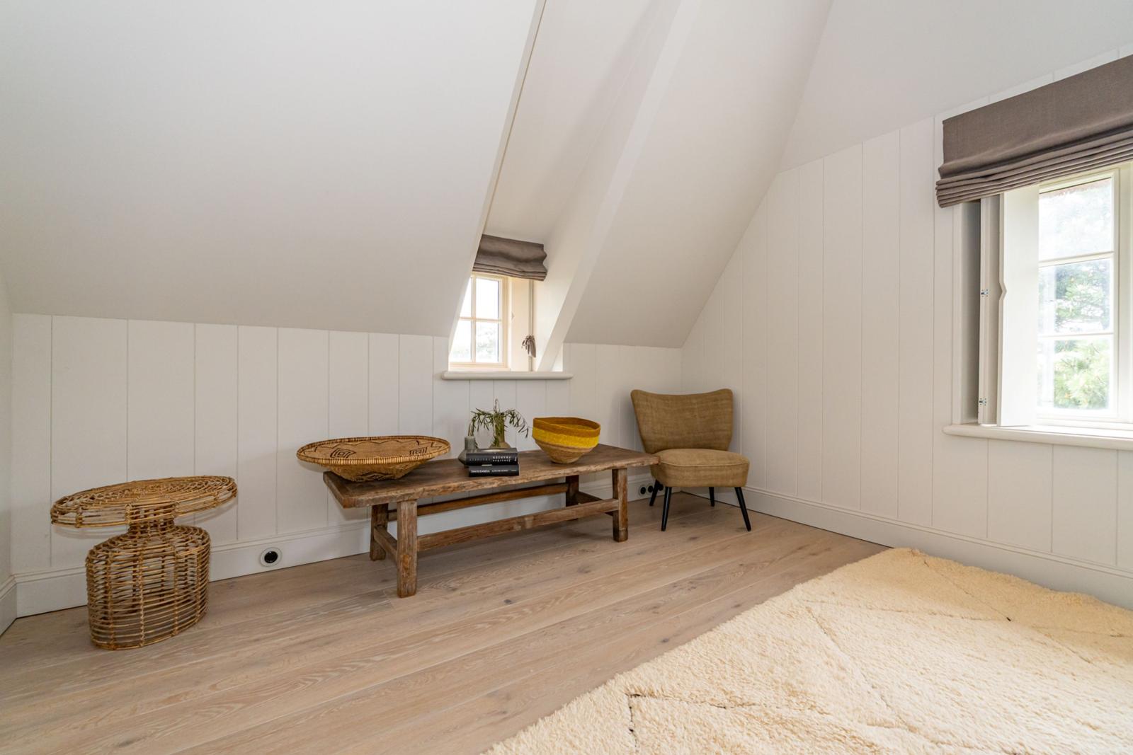 Ein Zimmer mit schrägen Wänden, Holzboden und zwei Fenstern. Möbel aus Holz und Rattan.