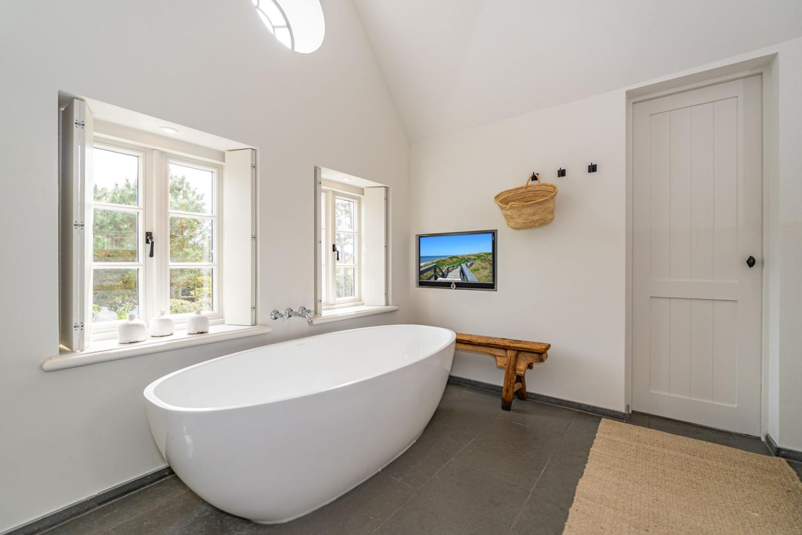 Bad mit freistehender Badewanne, Fenstern und Fernseher an der Wand.