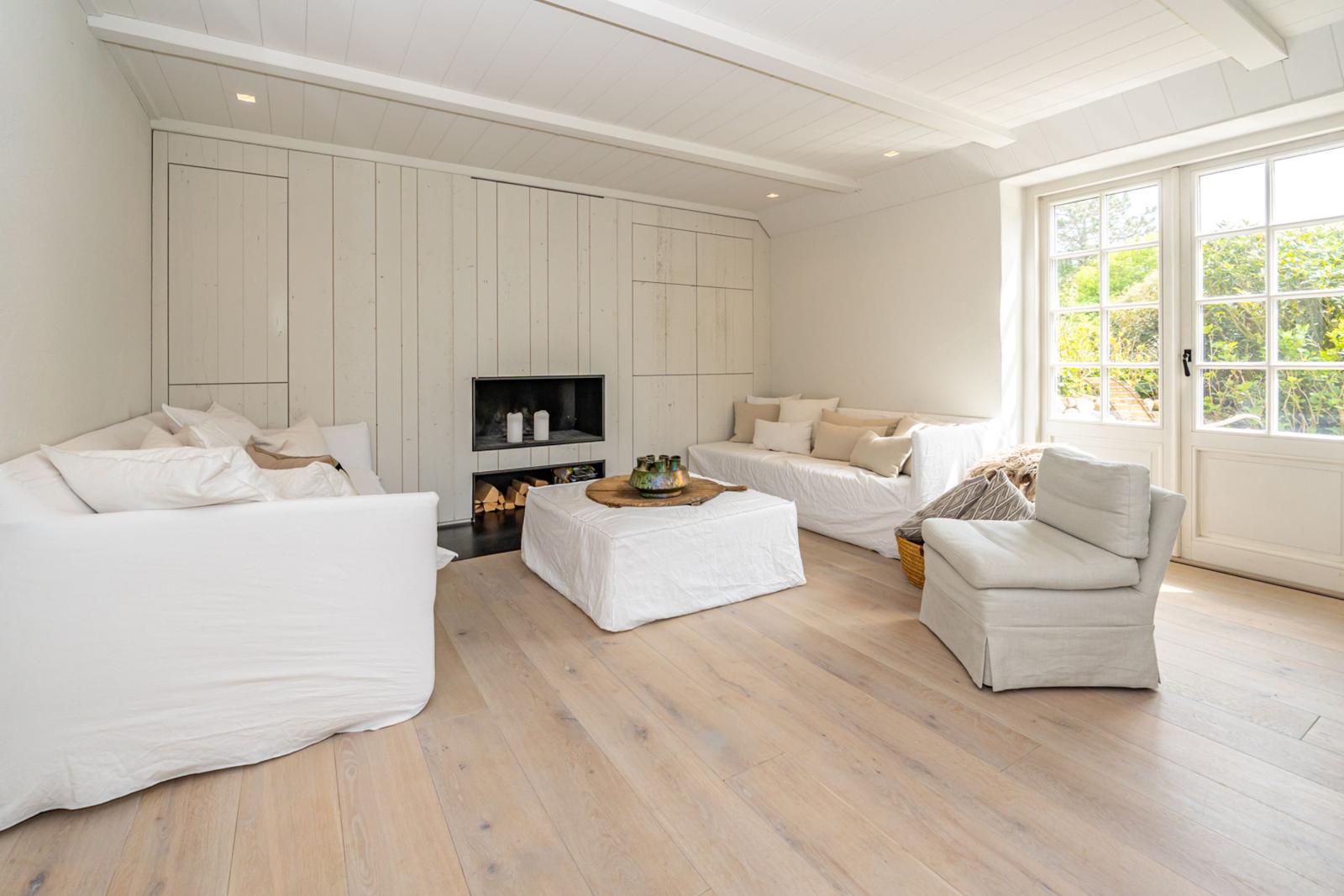 Wohnzimmer mit weißem Sofa, Kamin und Holzboden. Große Fenster mit Blick auf Grün.