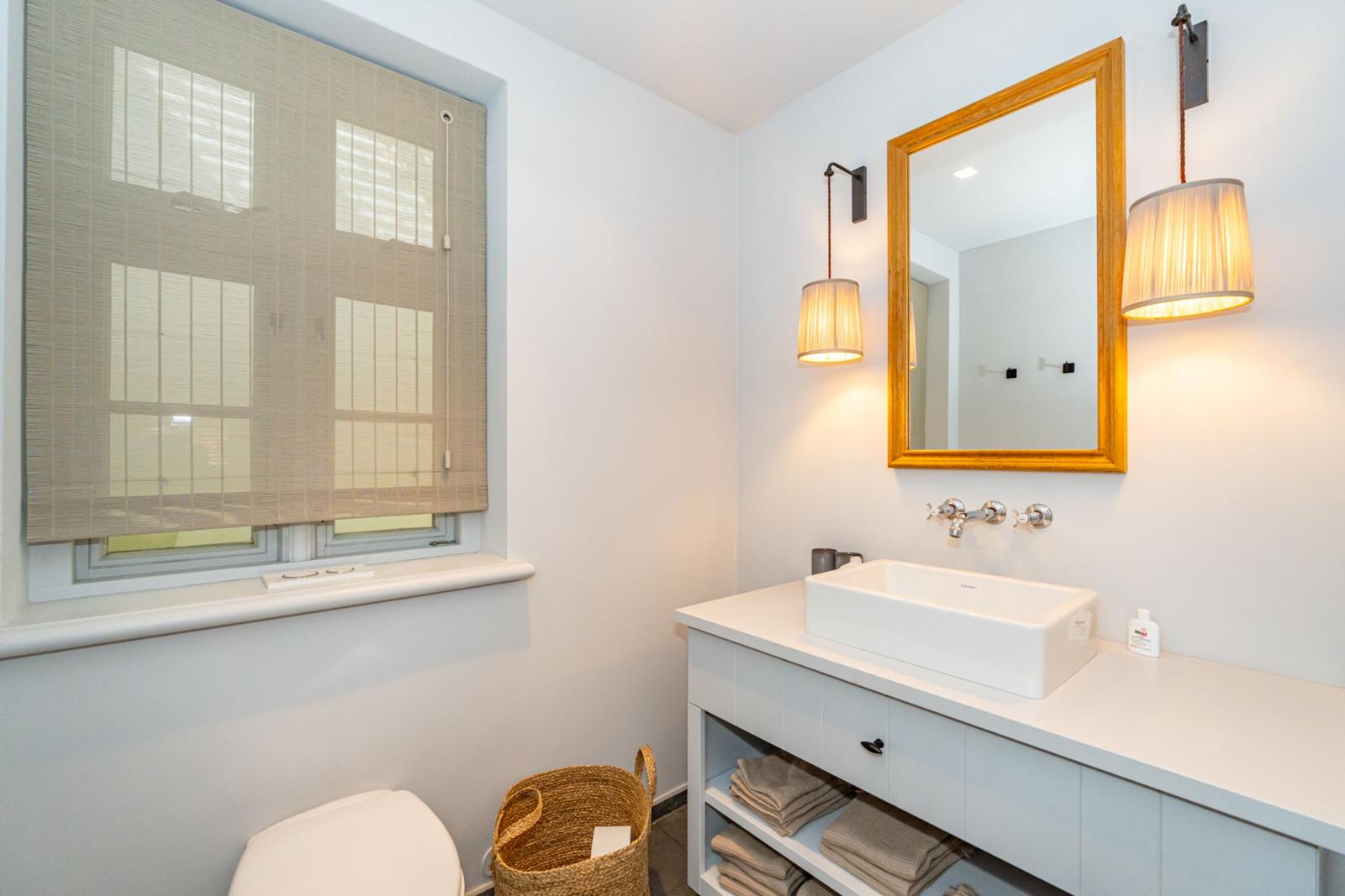 Badezimmer mit Waschbecken, Spiegel, Toilette und Fenster mit Jalousie.