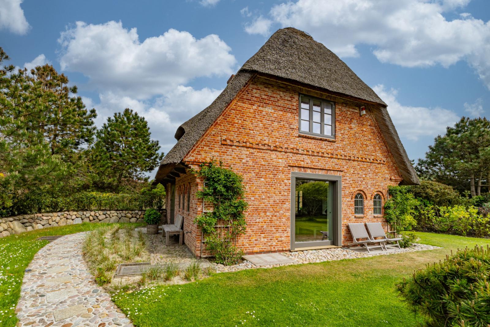 Ein rotes Backsteingebäude mit Strohdach und Gartenweg. Zwei Liegestühle stehen vor der Tür.