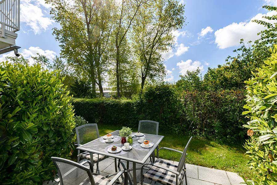 Terrasse mit Tisch und Stühlen, umgeben von grünen Hecken und Bäumen unter blauem Himmel.