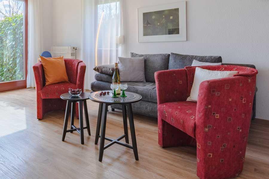 Wohnzimmer mit grauem Sofa, zwei roten Sesseln und zwei Tischen. Durch Fenster blickt man auf Grün.