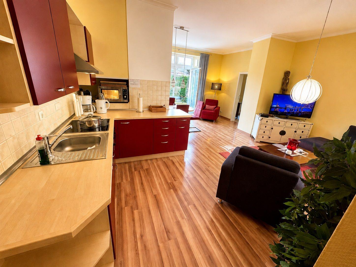 Küche mit roten Schränken und Wohnzimmer mit dunklem Sofa und Fernseher.