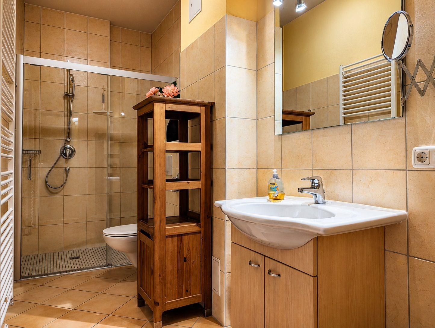 Bad mit Dusche, Waschbecken und Holzregal. Wandheizung und Spiegel sichtbar.