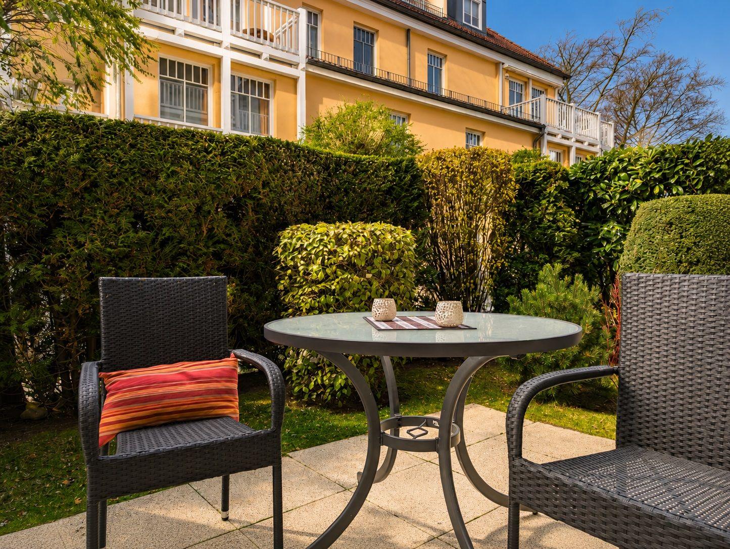 Außenterrasse mit Tisch und zwei Stühlen, umgeben von grünen Hecken und einem gelben Haus.