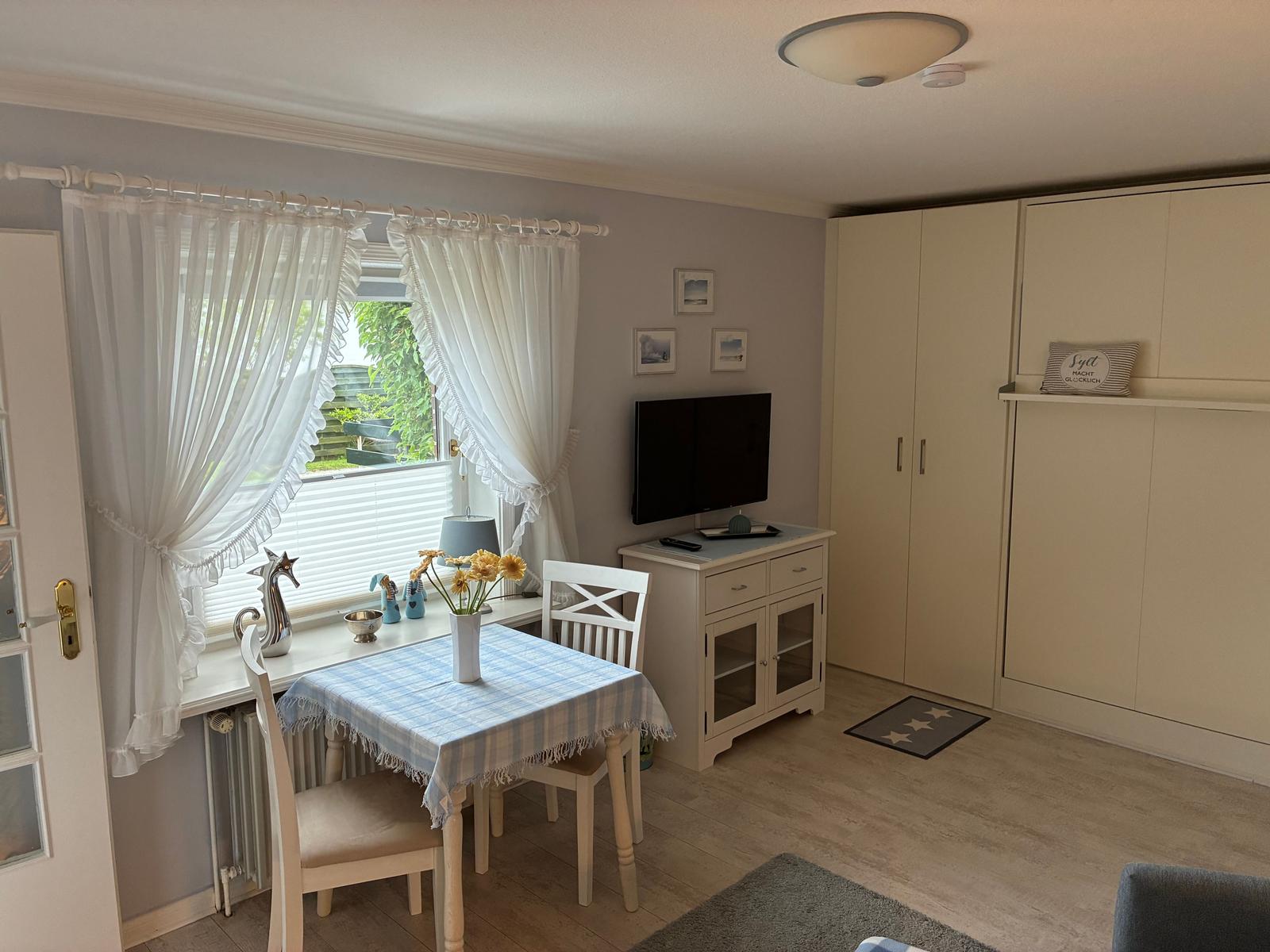 Ein Raum mit Esstisch, TV und Schränken. Fenster mit weißen Vorhängen.