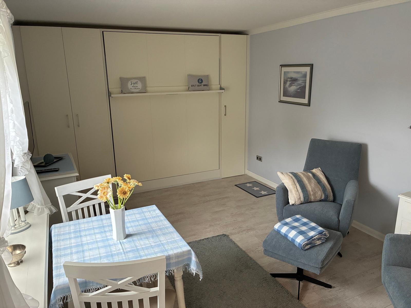 Ein Raum mit Essbereich, Sessel und Schrank. Tisch mit Blumen und Teppich.