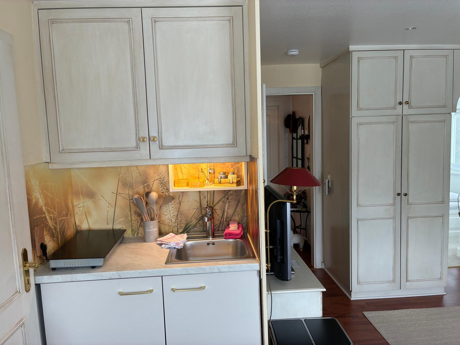 Küche mit Unterschrank, Spüle und Hängeschränken. Im Hintergrund steht ein Kleiderschrank.