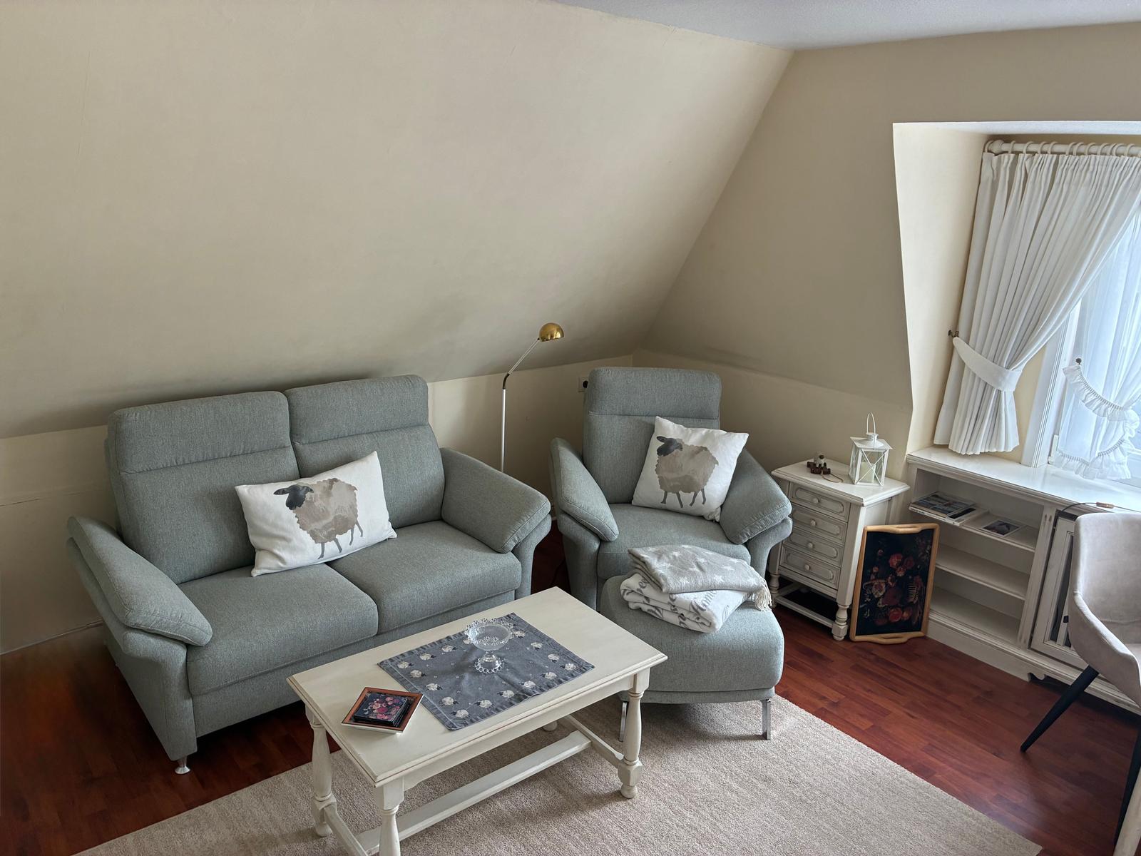 Ein Wohnzimmer mit grauem Sofa, Sessel und weißem Couchtisch. Fenster mit weißen Vorhängen.