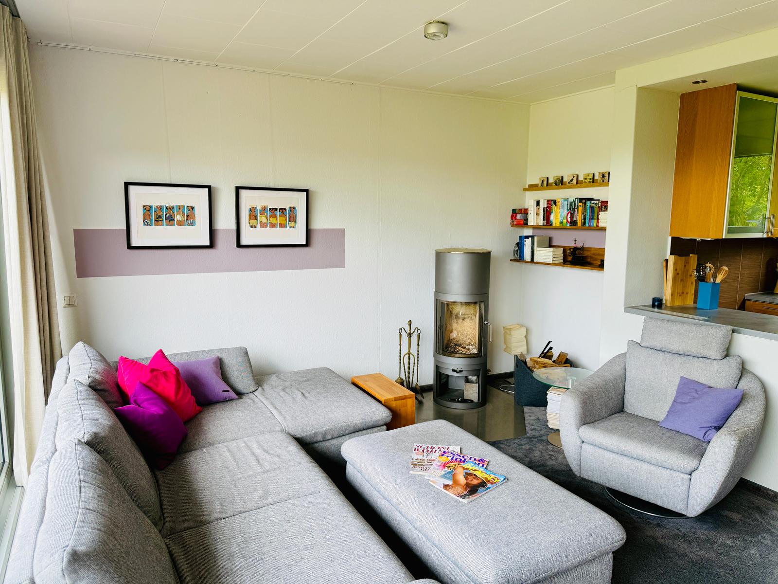 Wohnzimmer mit grauem Sofa, Kamin und Bücherregal. Küche im Hintergrund.
