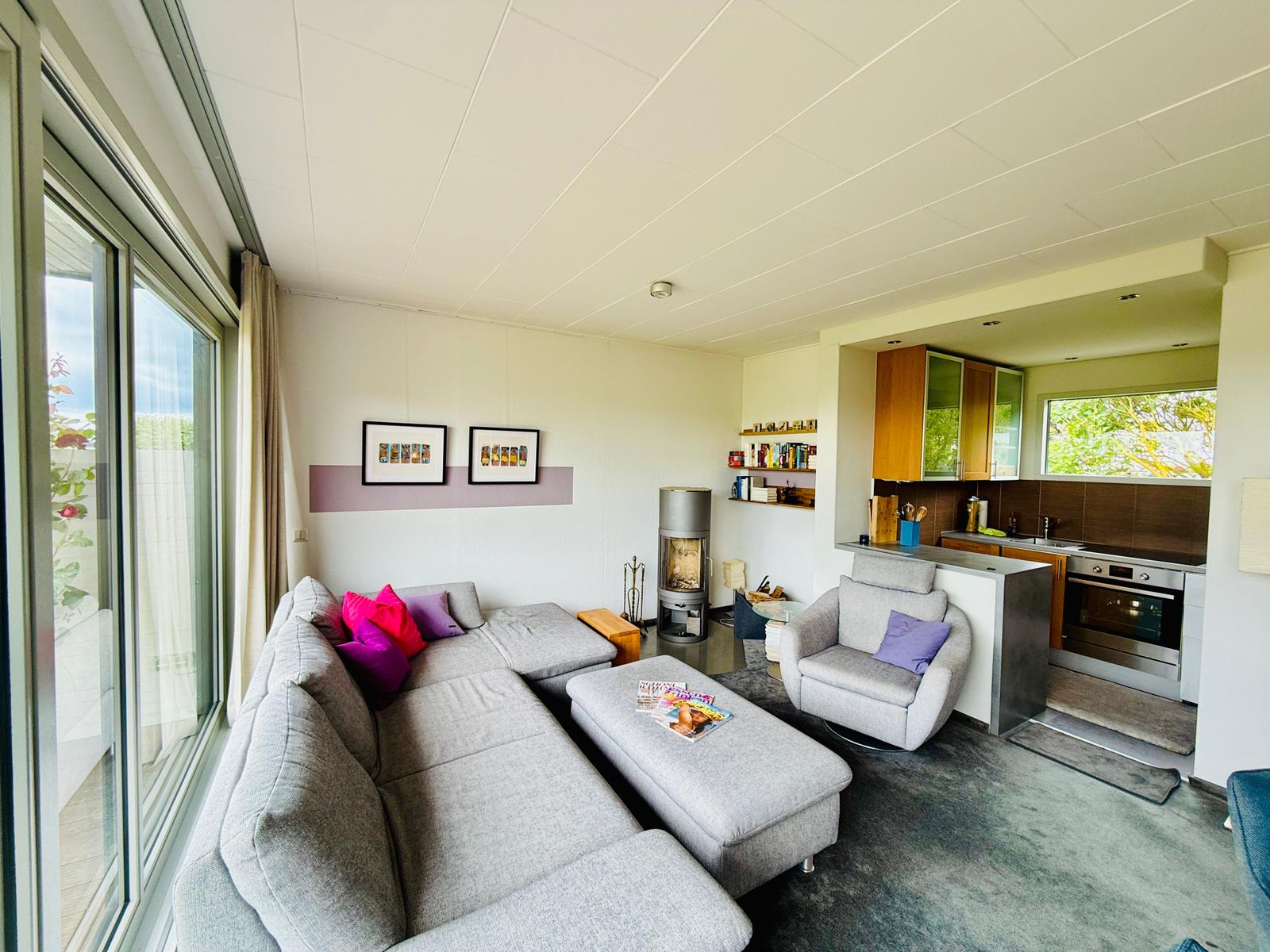 Wohnzimmer mit Sofa, Sessel und Küchenbereich. Große Fenster und Holzelemente.