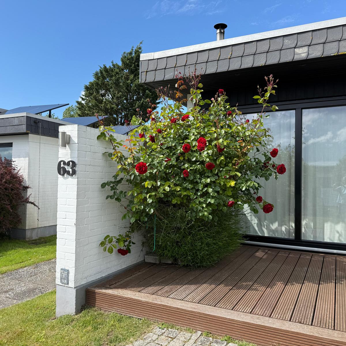 Haus mit roter Rosenhecke, Holzterrasse und weißem Mauerabschnitt mit Hausnummer 63.